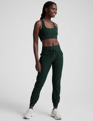 model wearing a green scoop neck racerback long line sports bra and joggers against a plain background.