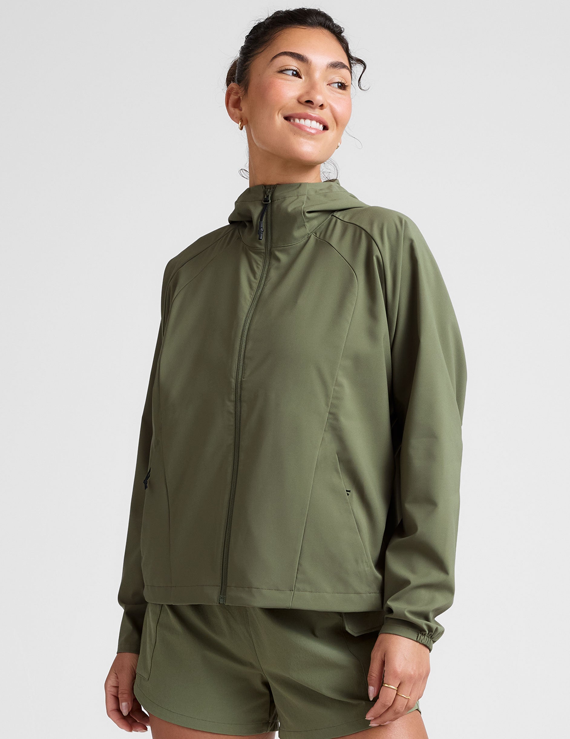model is wearing an olive green zip-up jacket with side pockets and a hood on a white background.