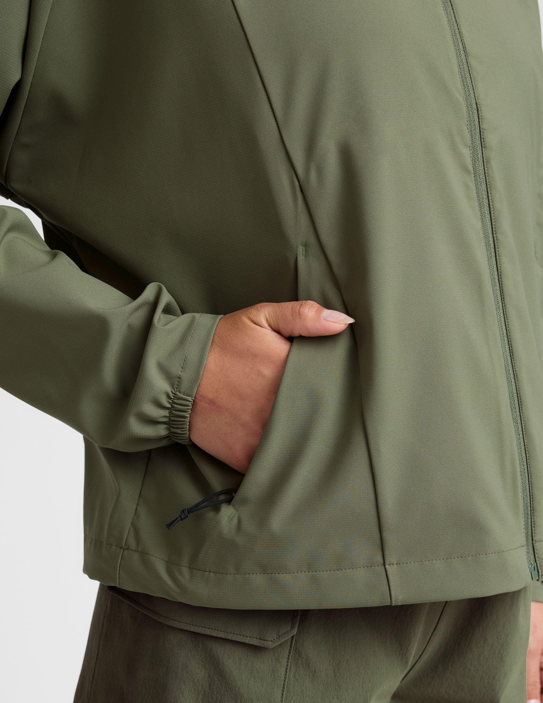 model is wearing an olive green zip-up jacket with side pockets and a hood on a white background.