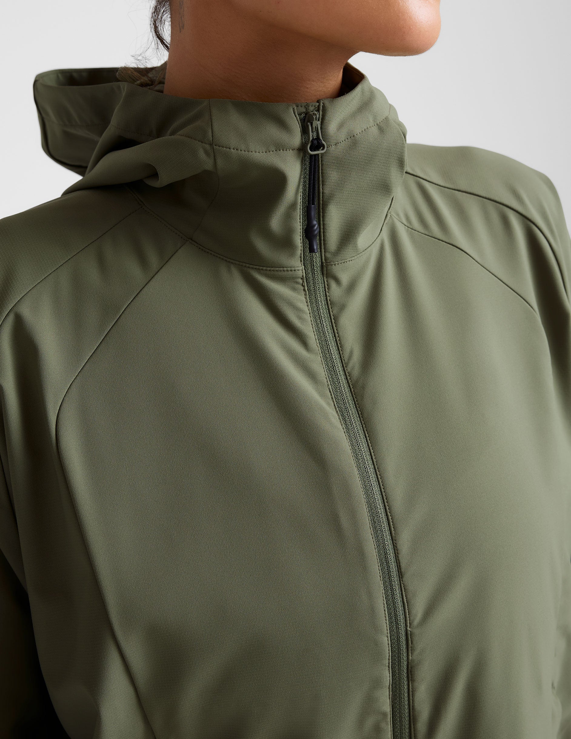 model is wearing an olive green zip-up jacket with side pockets and a hood on a white background.