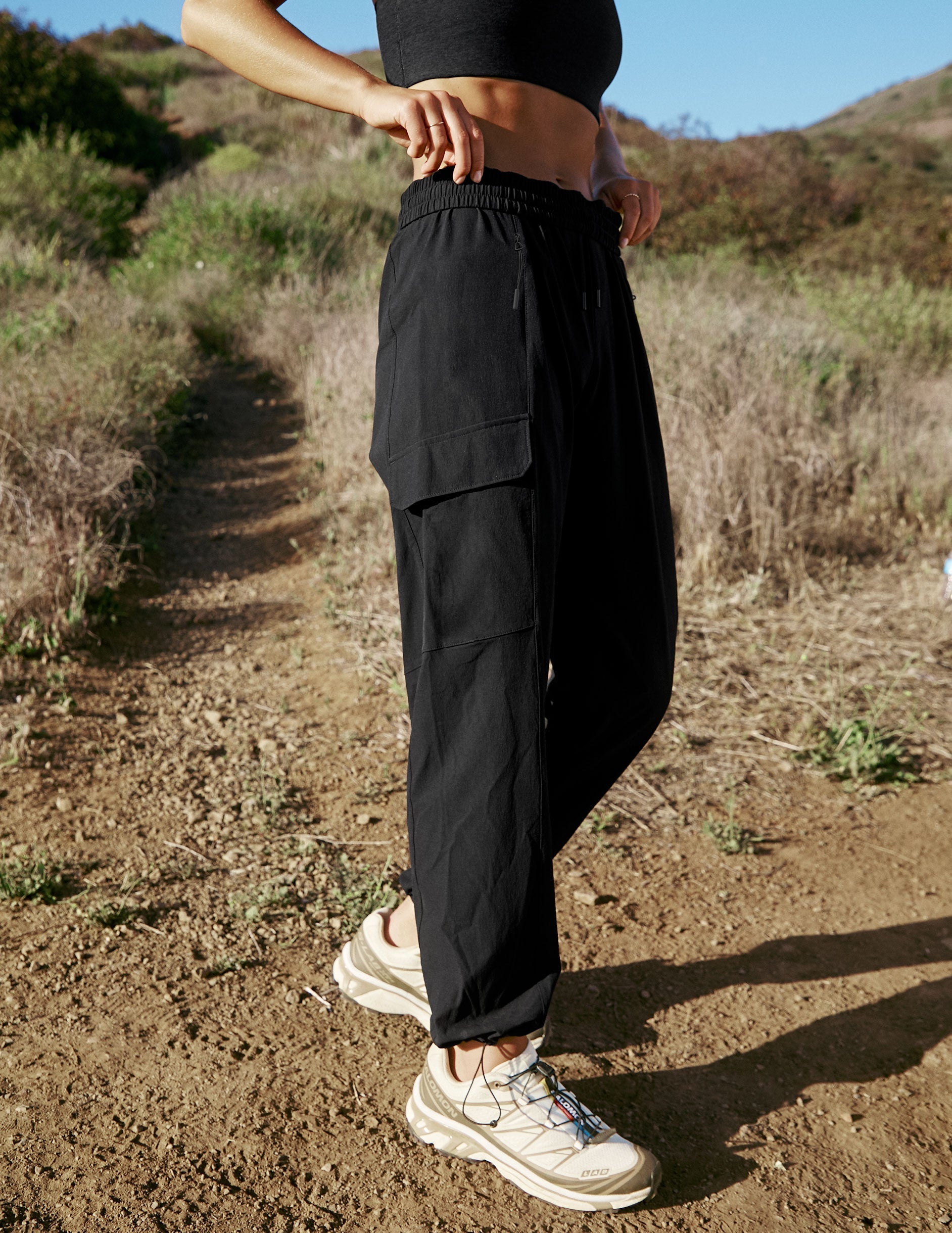 model wearing a black sports bra and black cargo style pants hiking outdoors with mountains in the background.
