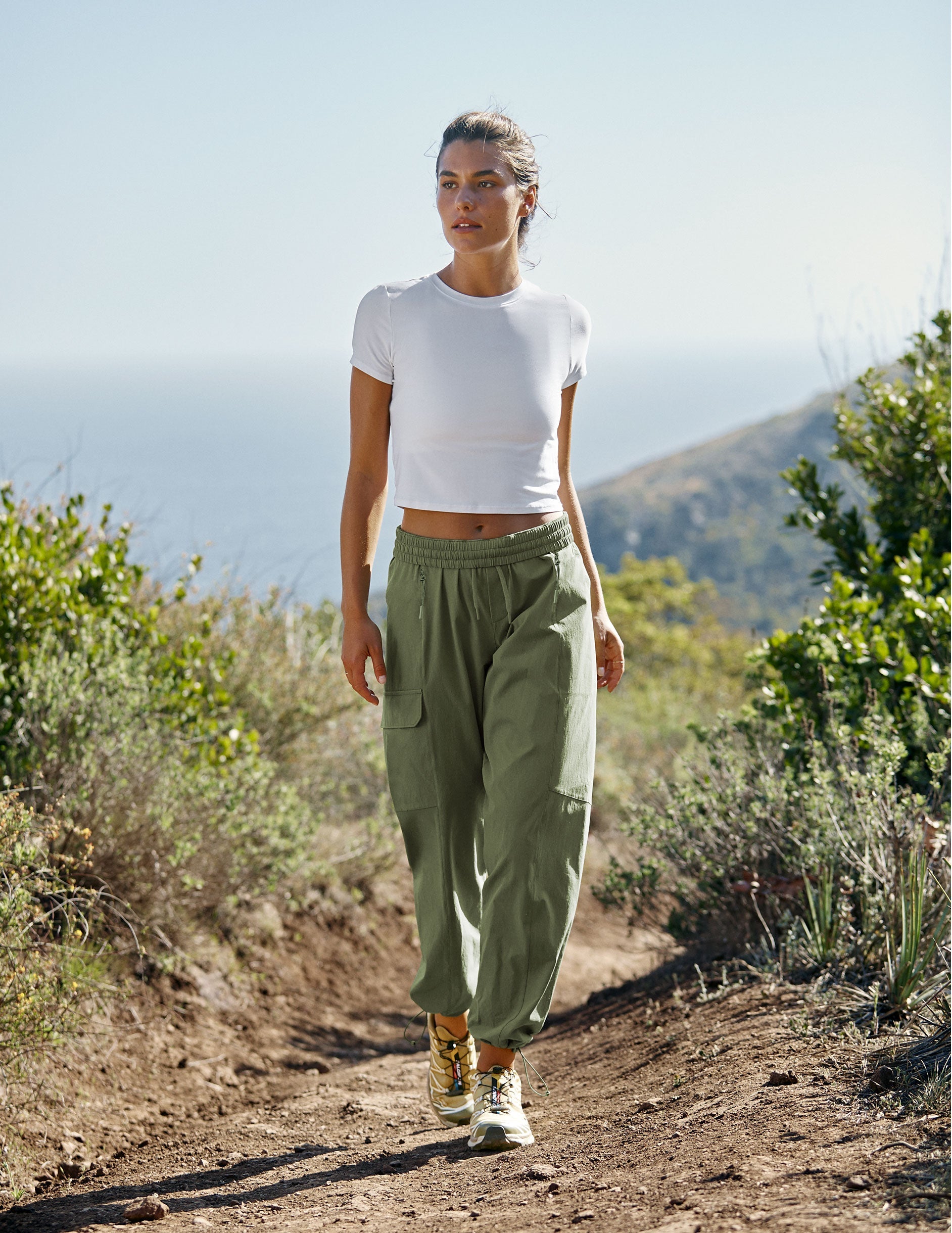 model wearing a white t-shirt and green cargo style pants walking on a trail in nature wearing a white top and green pants.