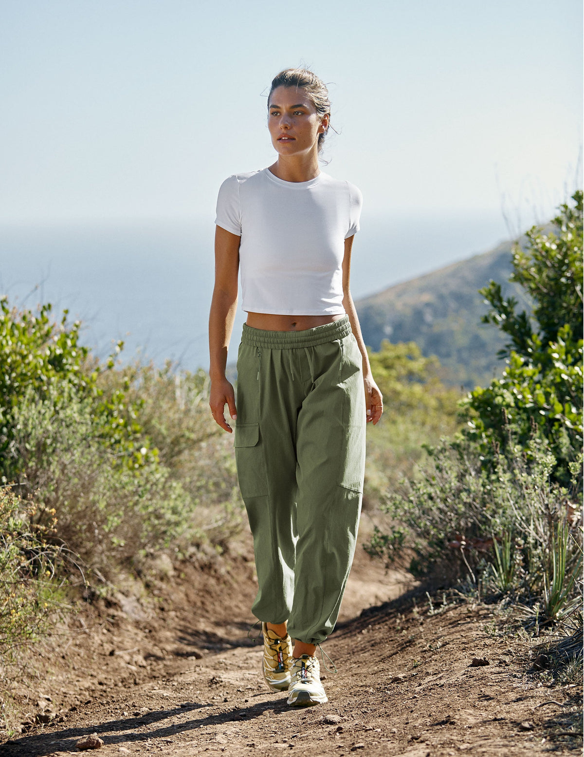 model wearing a white t-shirt and green cargo style pants walking on a trail in nature wearing a white top and green pants.