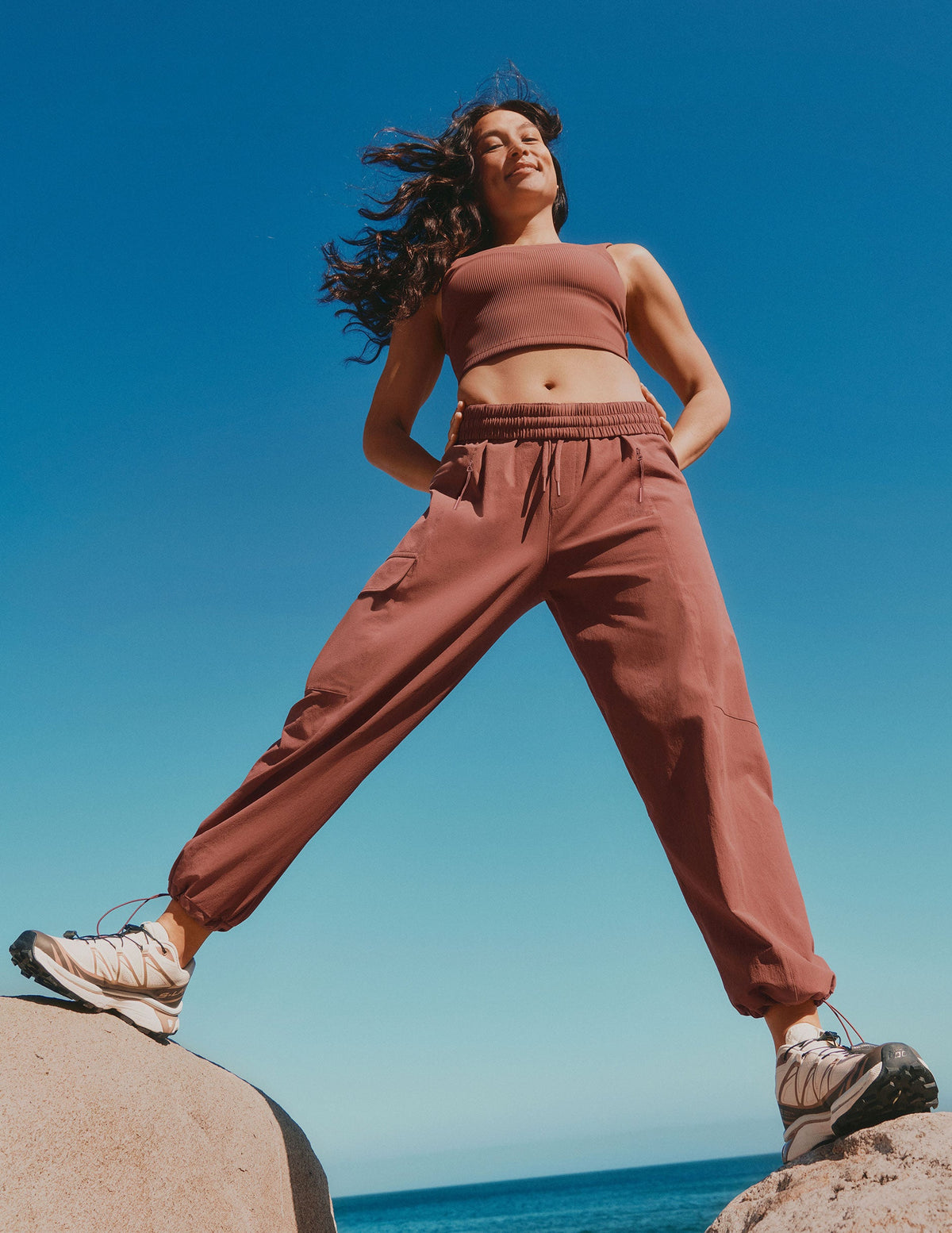 model in brown ribbed cropped tank and brown peak pants standing on a rock with blue sky and ocean background.