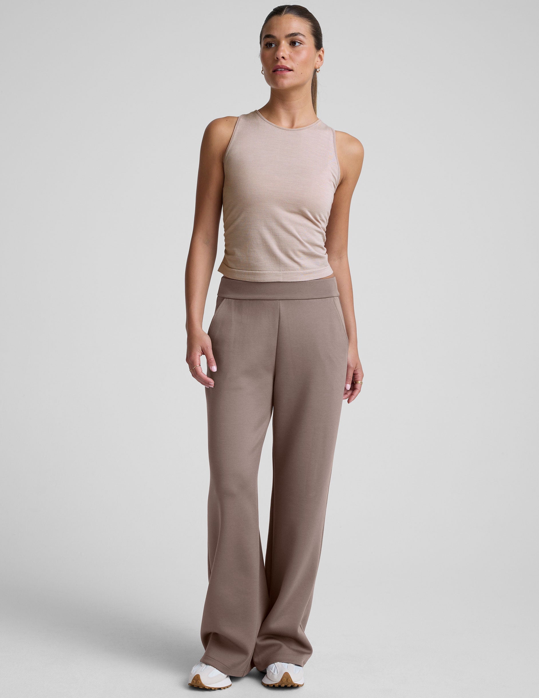 model is wearing a beige sleeveless top and brown pants against a plain background.