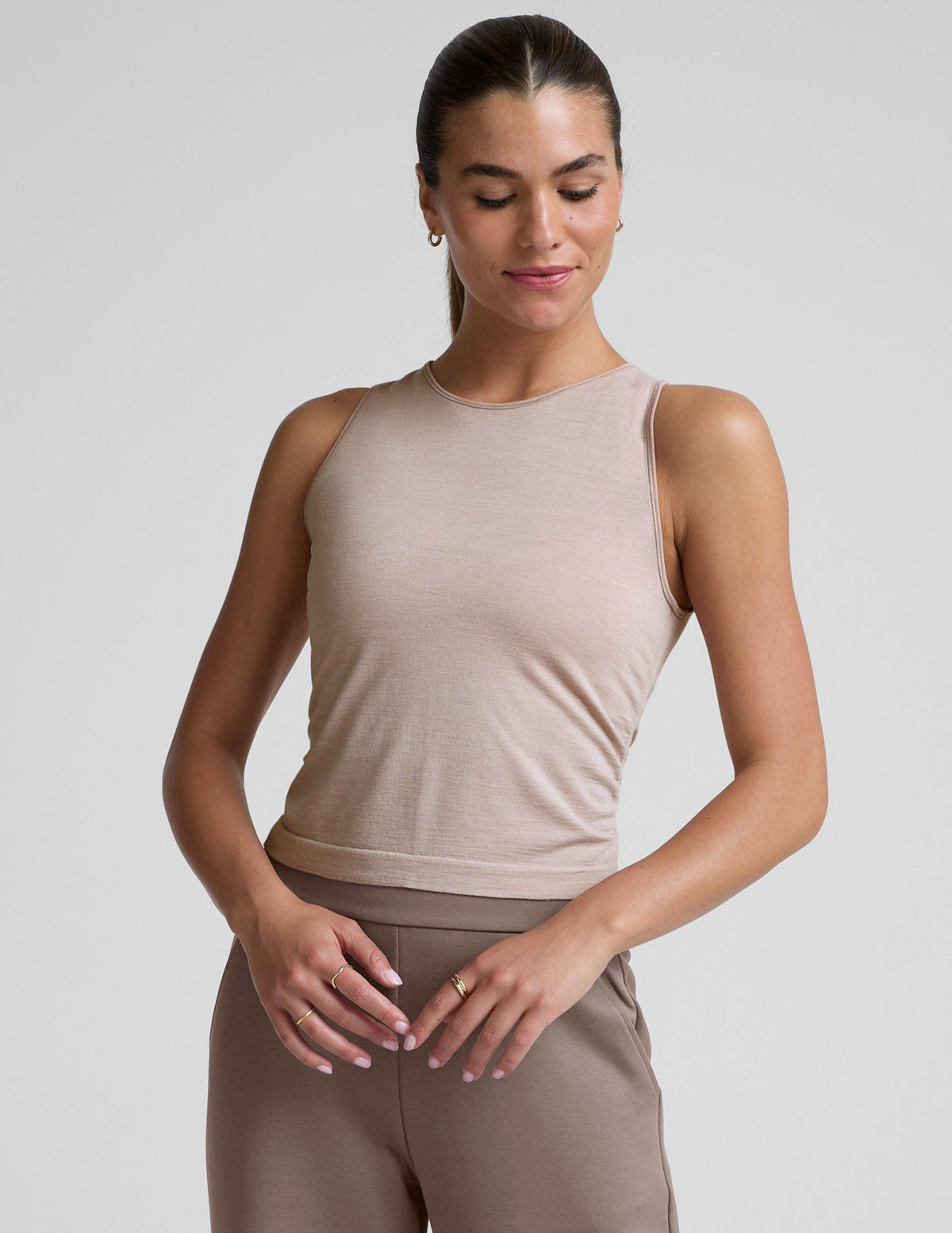 model is wearing a beige sleeveless top and brown pants against a plain background.
