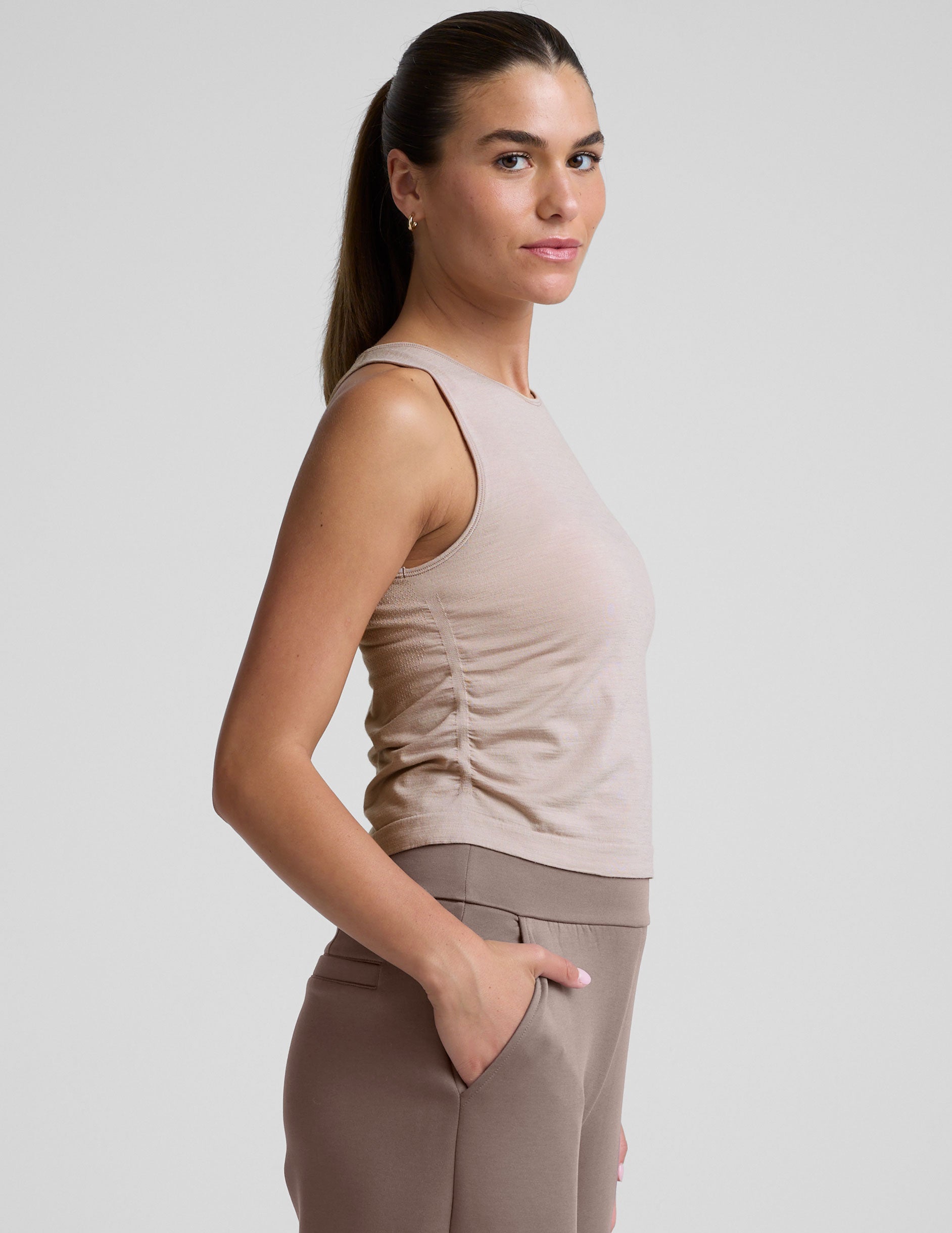 model is wearing a beige sleeveless top and brown pants against a plain background.