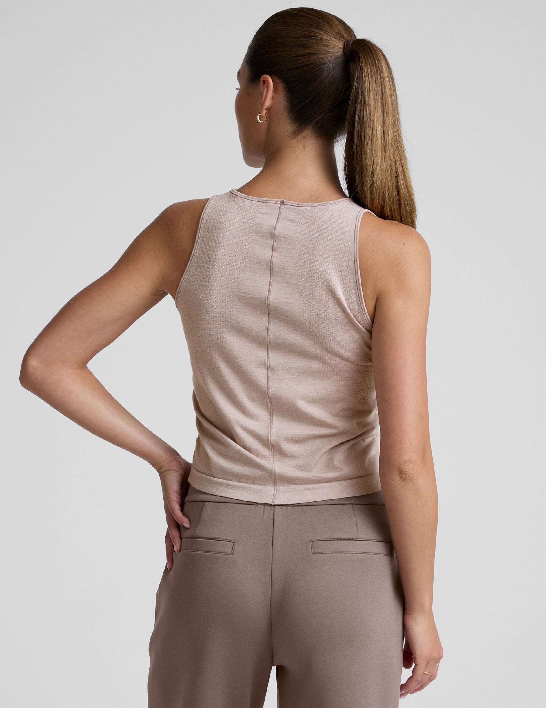 model is wearing a beige sleeveless top and brown pants against a plain background.