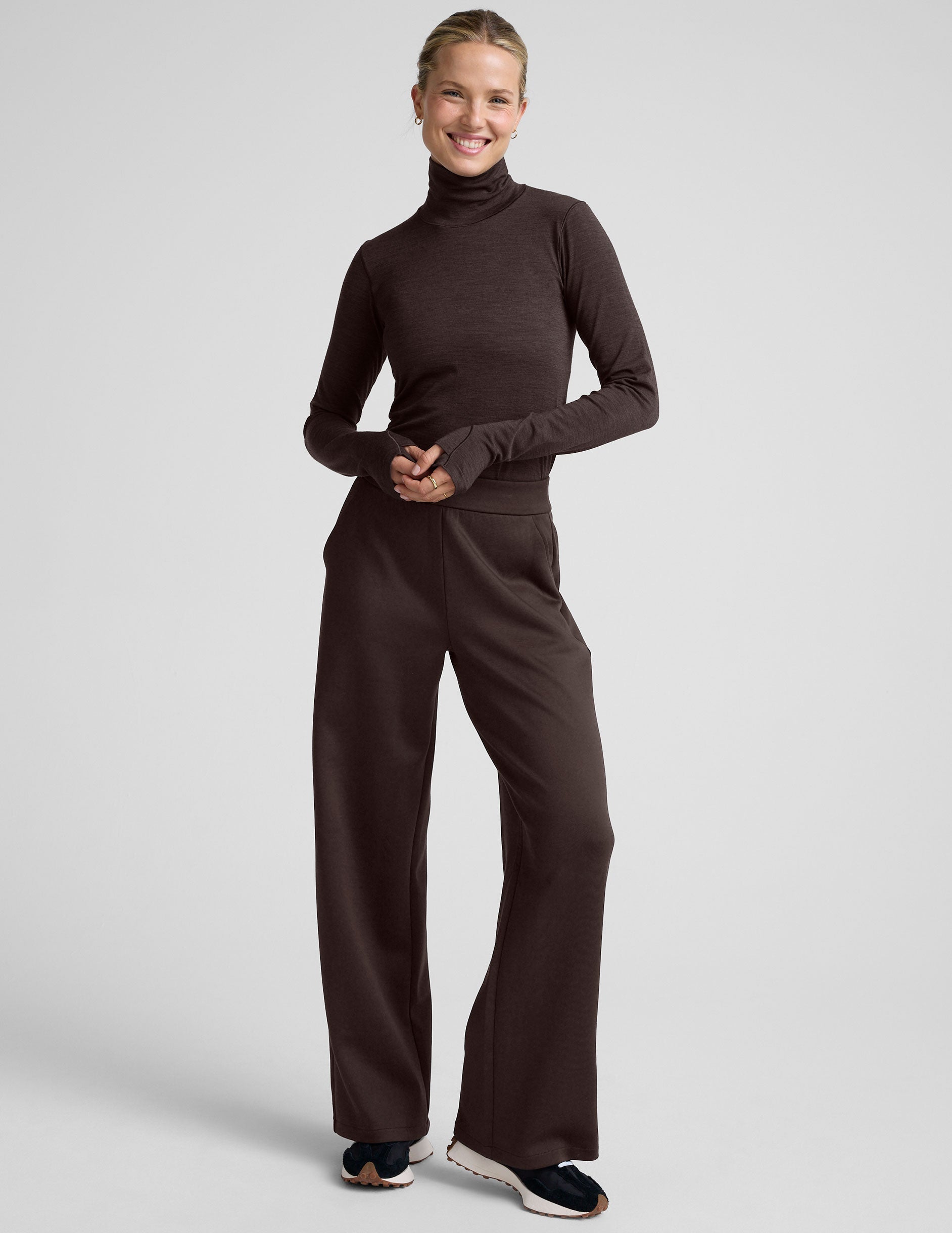 woman wearing a brown turtleneck and pants on a light gray background.