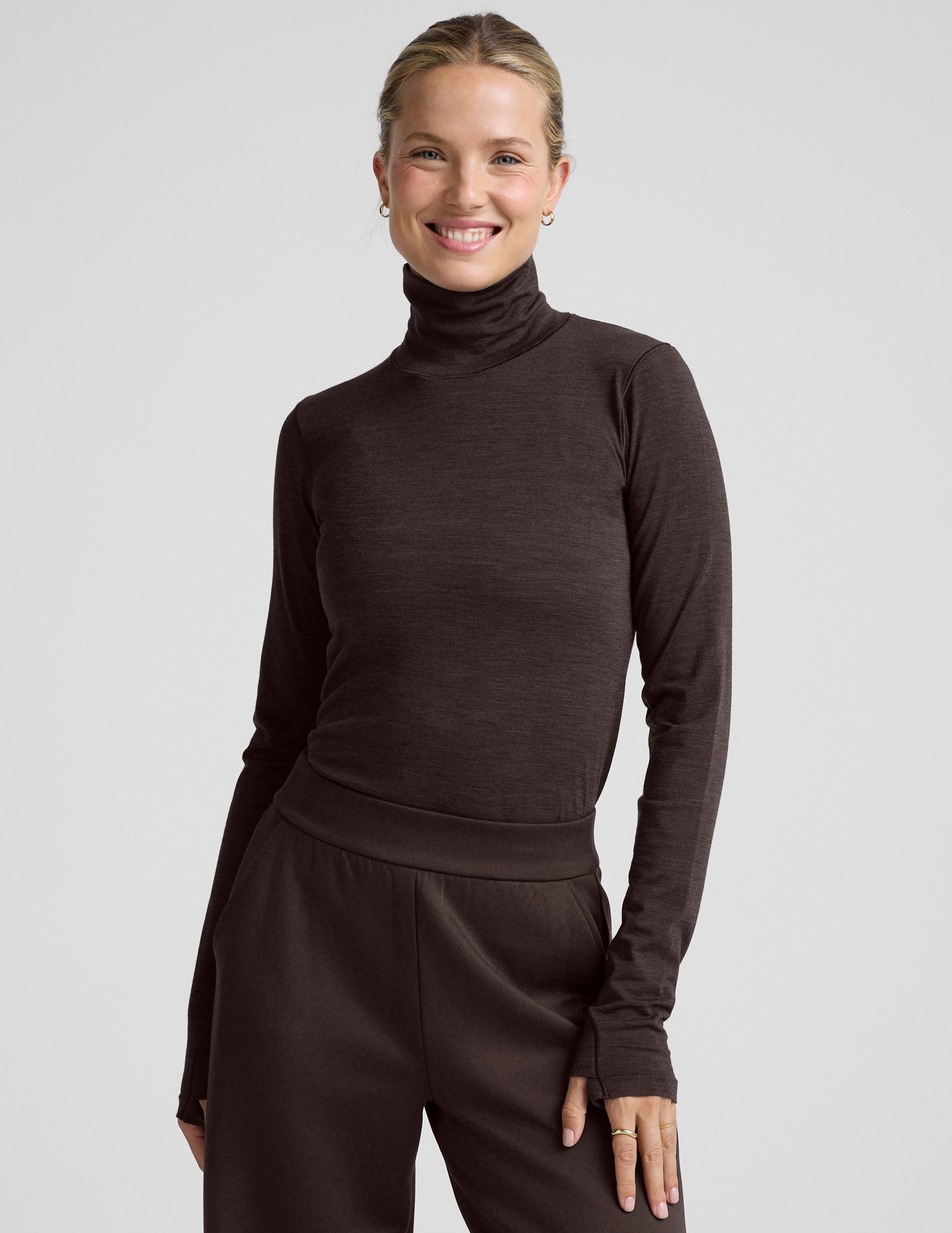 woman wearing a brown turtleneck and pants on a light gray background.