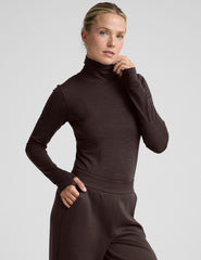 woman wearing a brown turtleneck and pants on a light gray background.