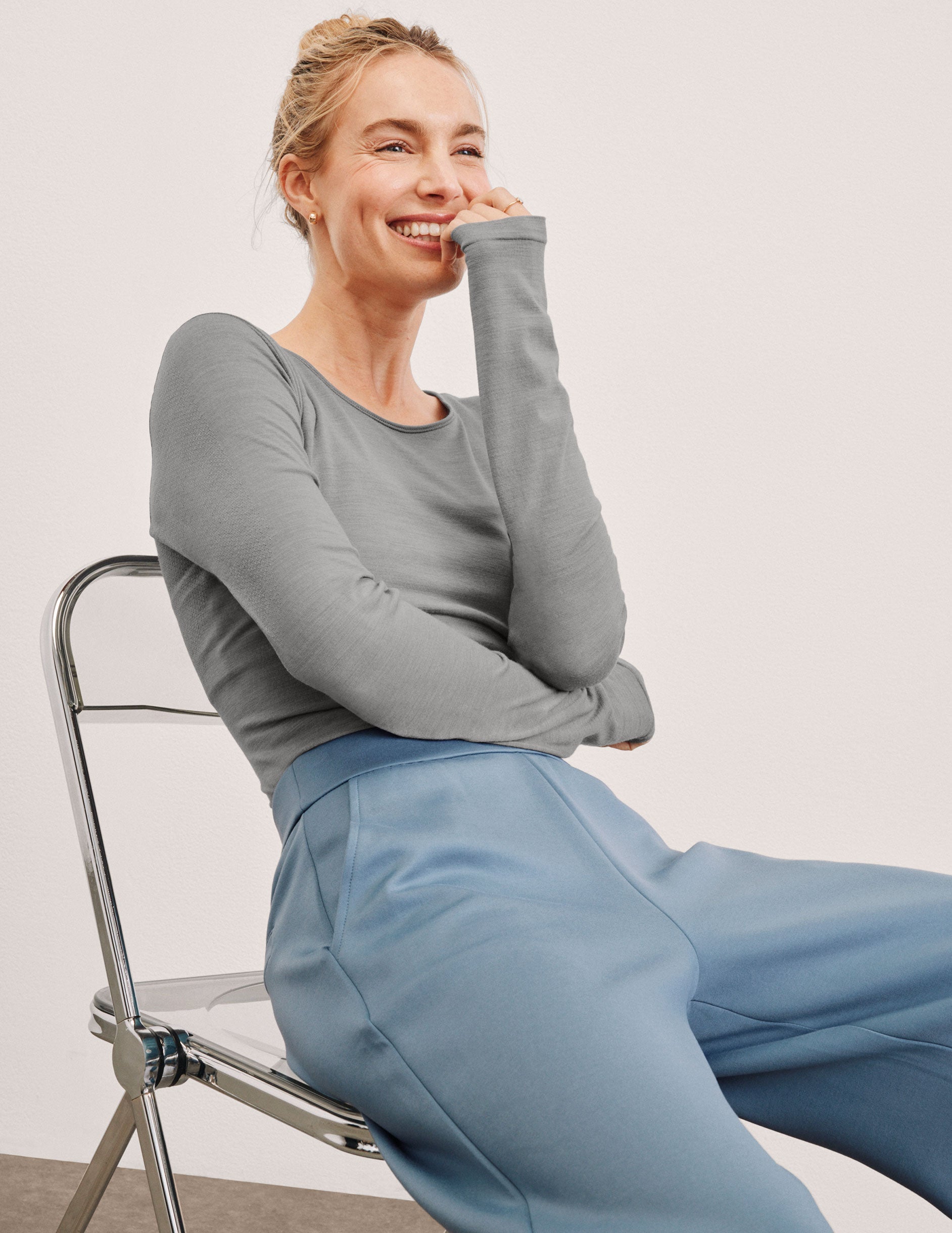 model sitting on a chair wearing a gray long-sleeve crewneck top and blue pants against a plain background.